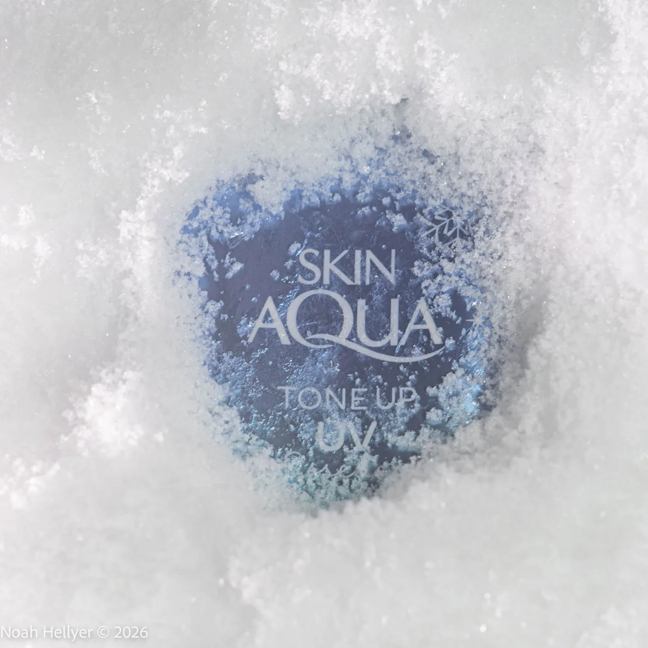 A blue Skin Aqua sunscreen tube pressed into fresh snow, glowing from within.
