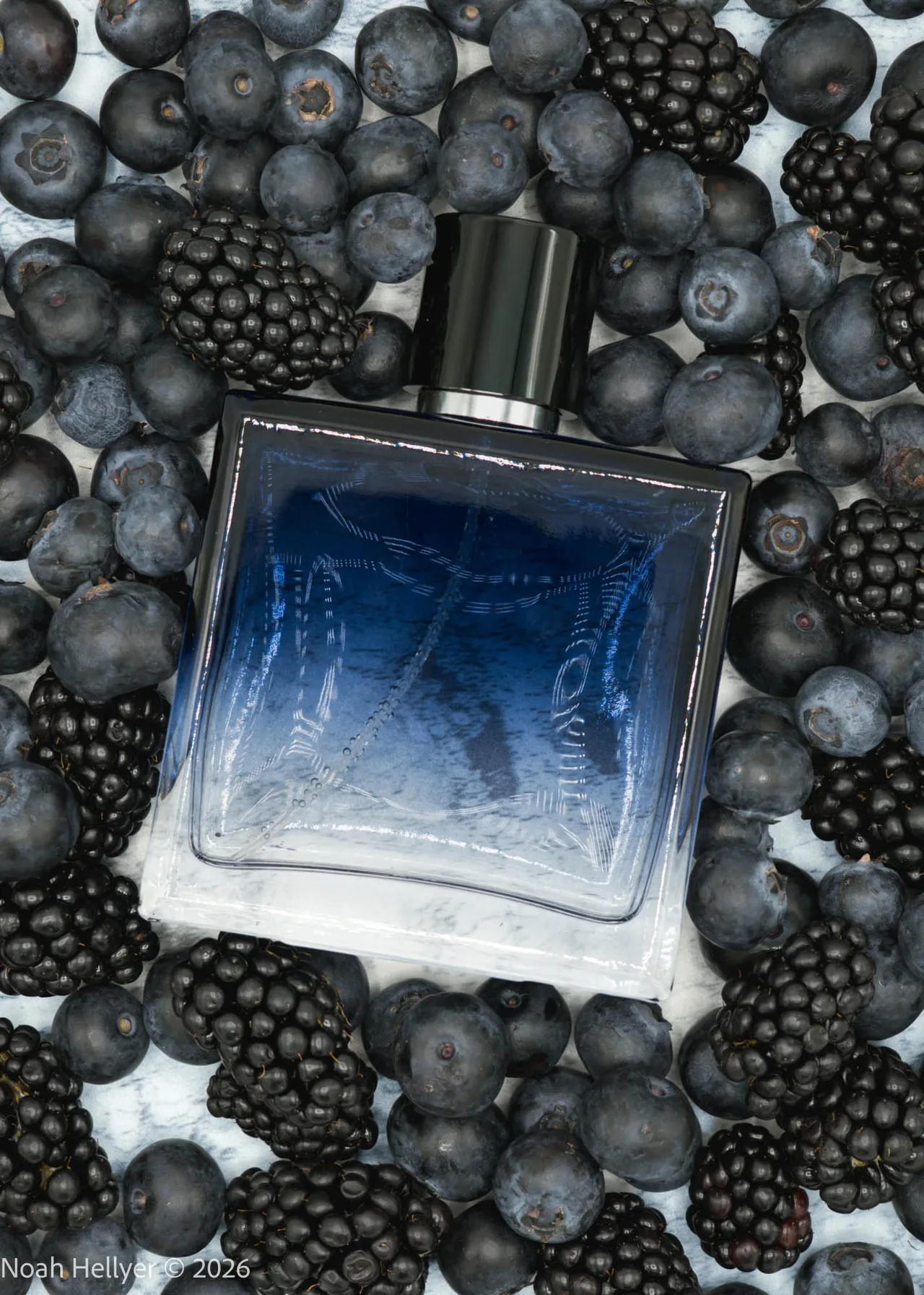 A clear blue perfume bottle lying in a bed of blueberries and blackberries, seen from above.