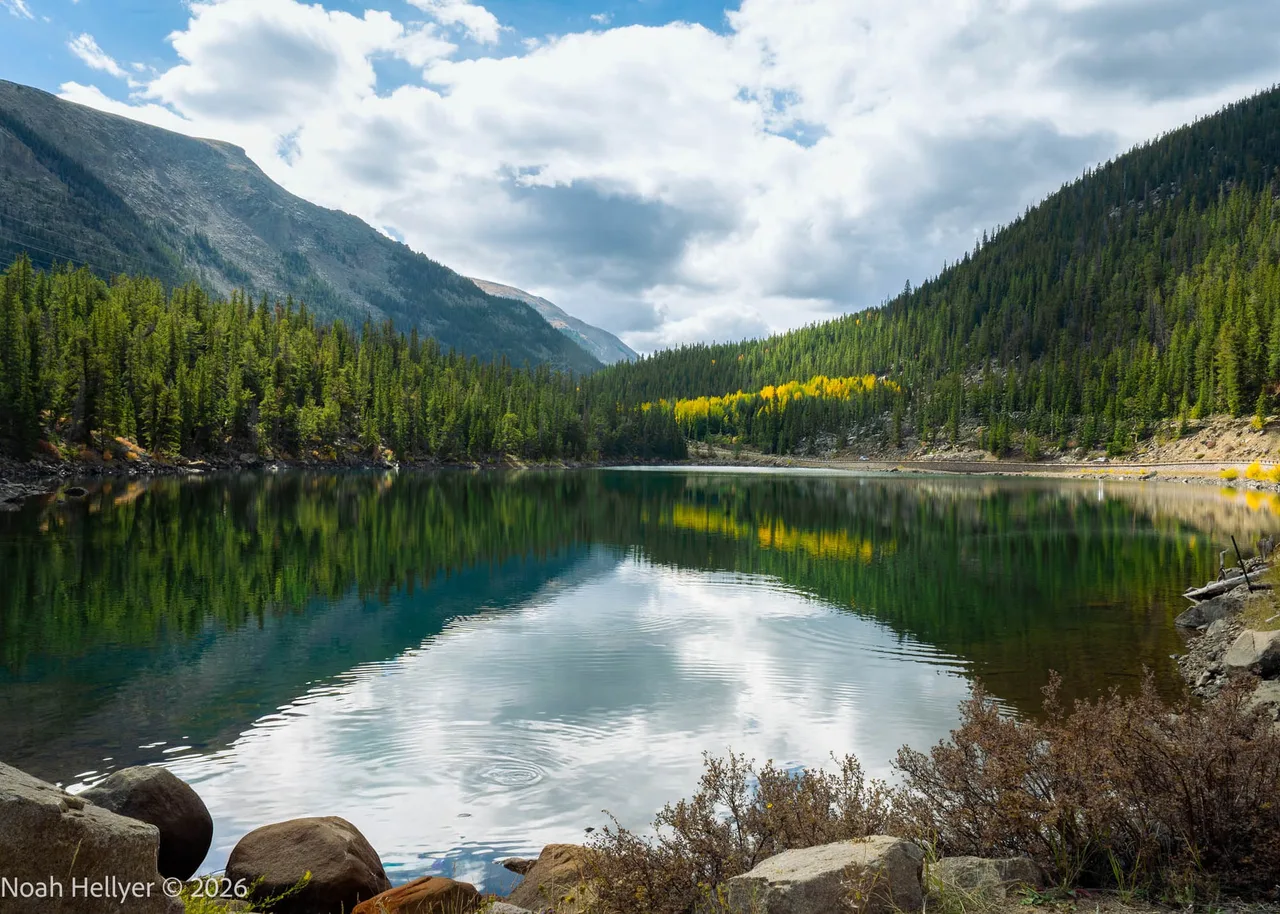 An alpine lake reflecting clouds and forested ridges, with boulders along the near shore.