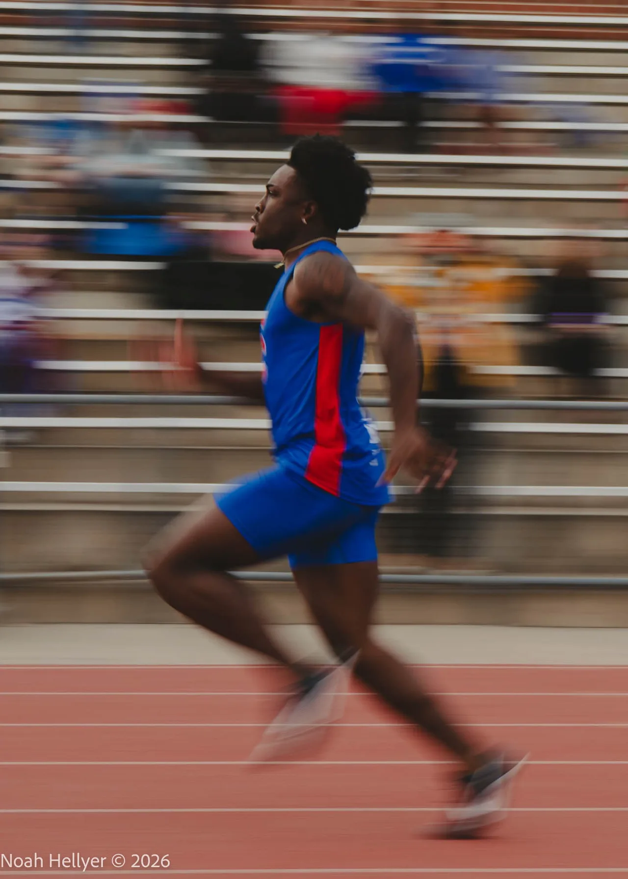A sprinter in a blue uniform mid-stride, the bleachers behind him streaked by a panning blur.