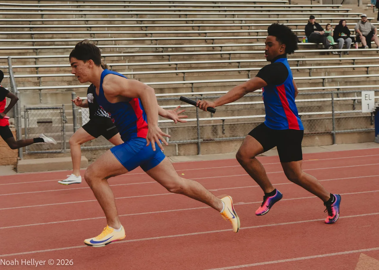 Two relay runners on a red track at the moment one hands the baton to the other.