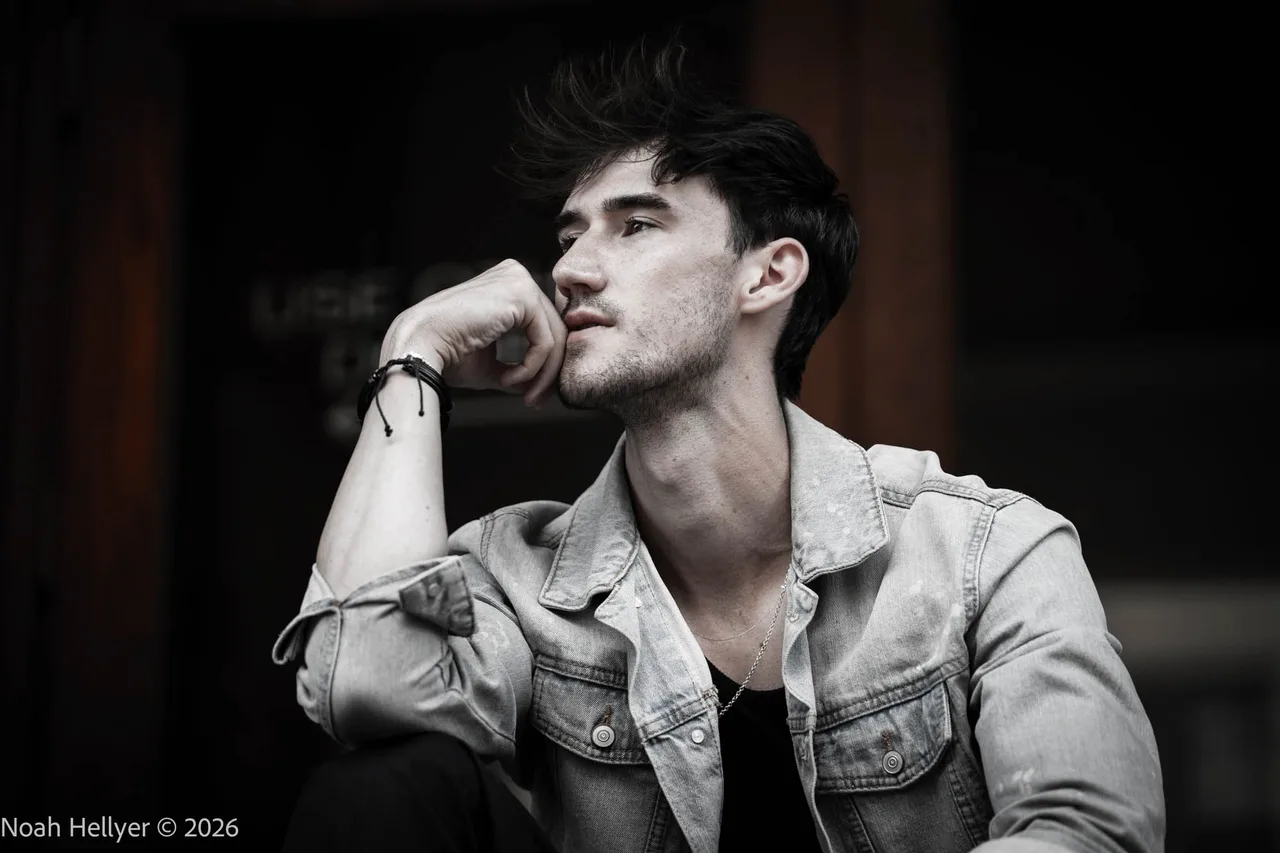 Black-and-white portrait of a young man in a denim jacket, chin resting on his hand.