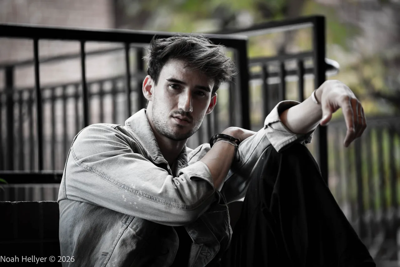 Black-and-white portrait of a man in a denim jacket sitting against an iron railing.