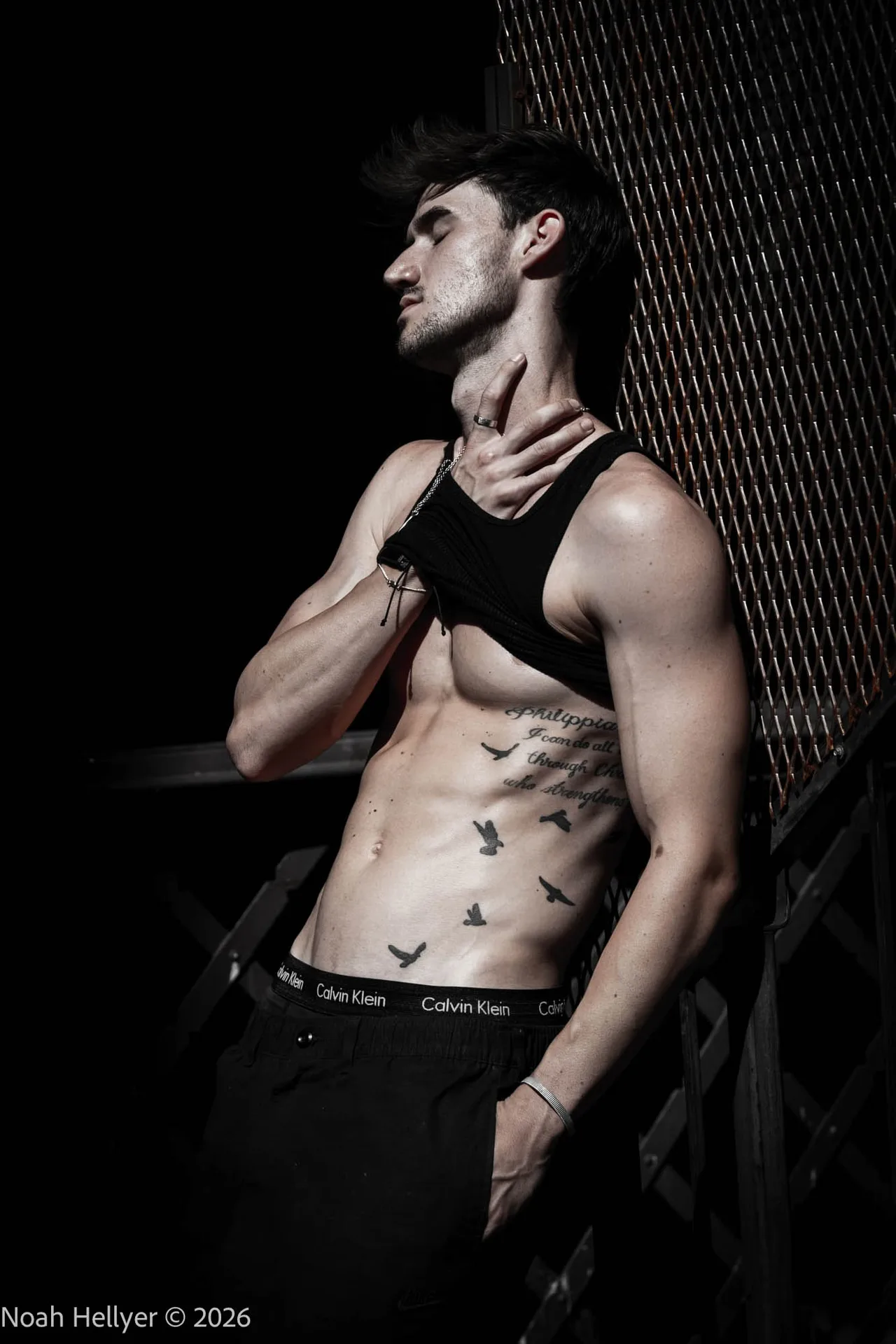 A shirtless man with a cropped tank top pulled up, head tilted back against a dark wall.