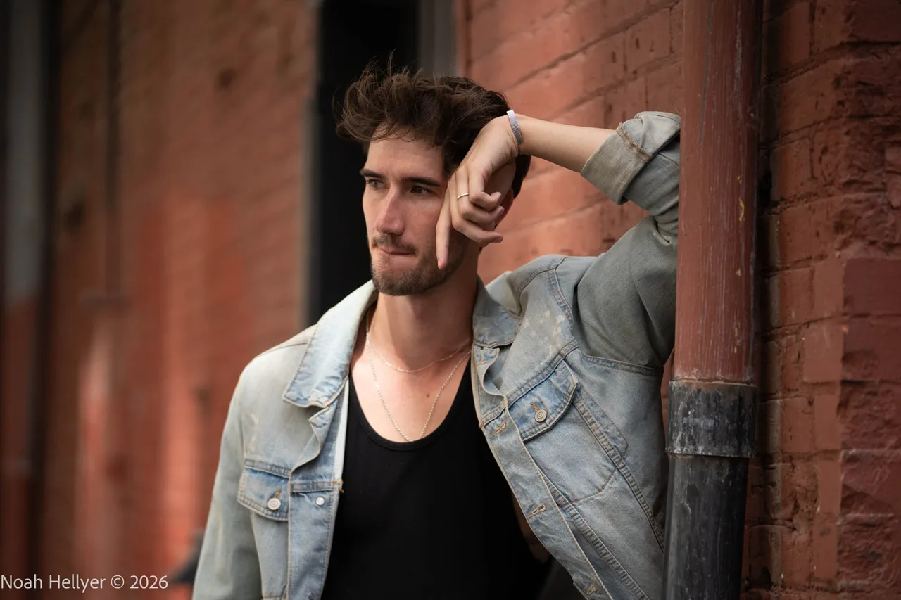 A man in a denim jacket leaning against a brick wall beside a steel pipe.