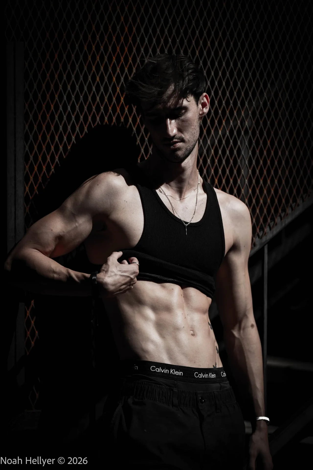 A man lifting the hem of his black tank top in a dark alley, lit by a single slice of skylight.
