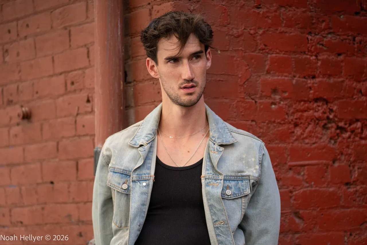 A man in a denim jacket and black tank top in front of a red brick wall.