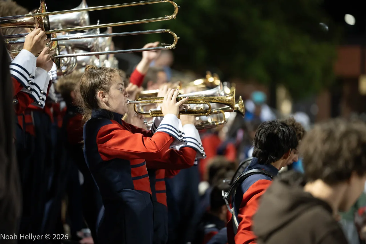 Brass players in red and black marching band uniforms playing under stadium lights.