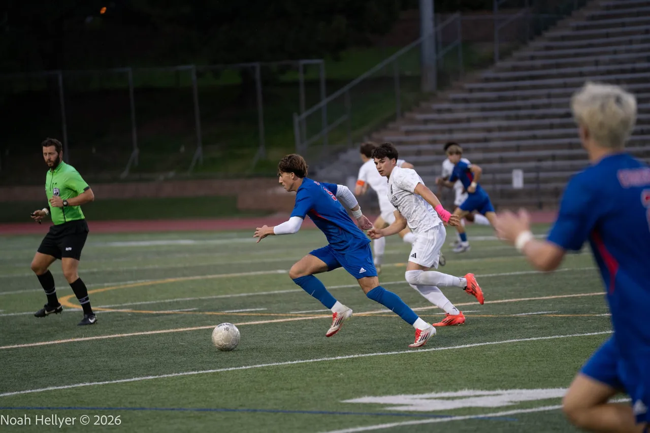 Varsity soccer players in blue and white chasing the ball across a lit field.