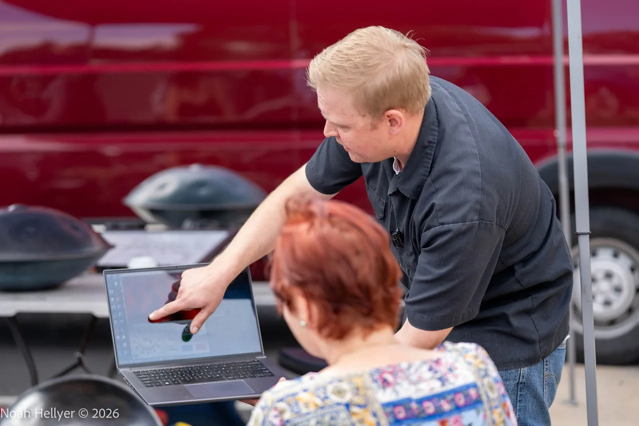 An instructor leaning over a table to show a student how to tune a steel pan, a handpan beside them.