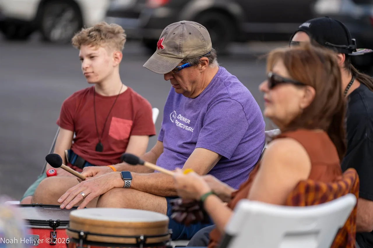 People seated in a circle playing hand drums in a parking lot.
