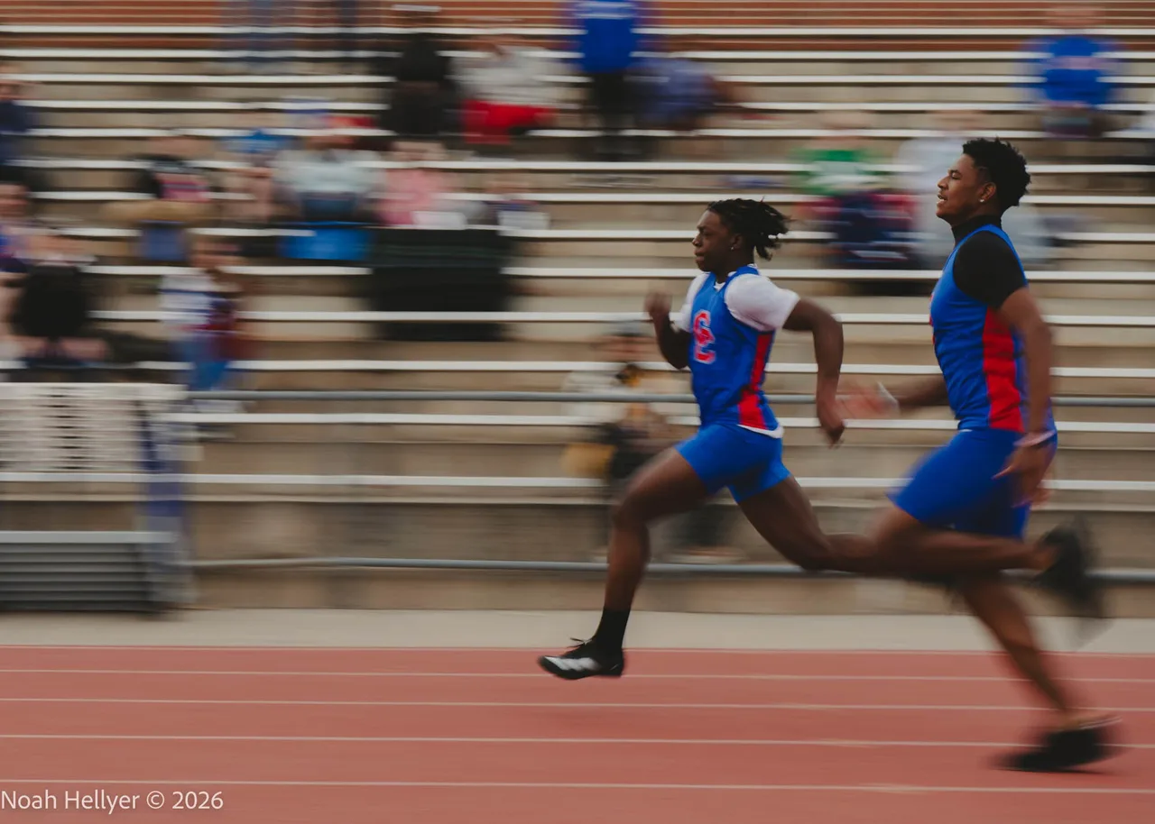 Two sprinters racing past a blurred grandstand on a red track.