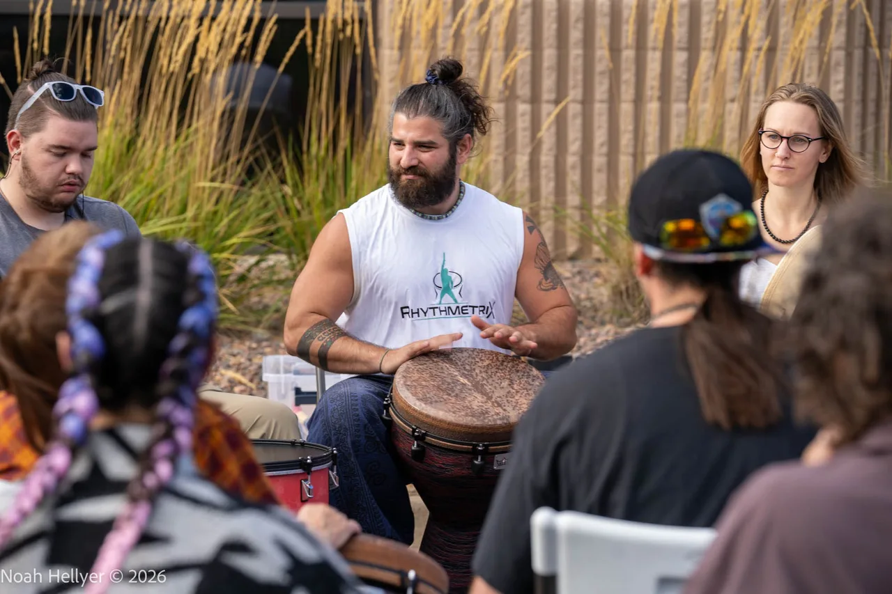 A bearded drummer leading a circle of seated players on djembes.